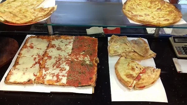 Il banco a vetri della pizzeria: teglie di pizza al taglio bianca e rossa appena tagliata, focacce farcite sulla carta e due focacce tonde sul ripiano di sopra.